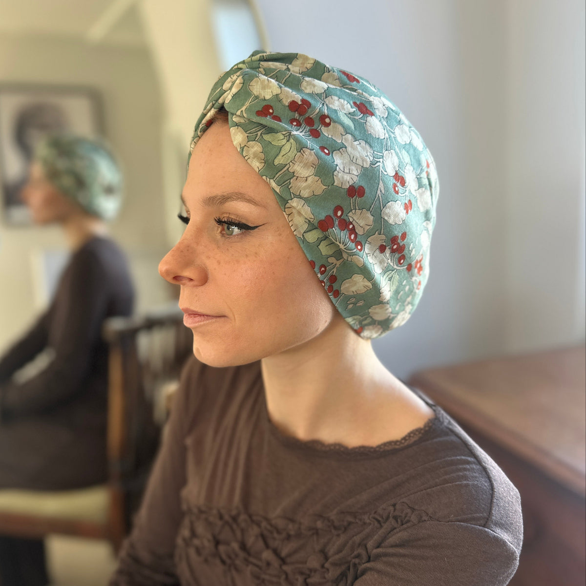 A woman in profile wearing a sage green fabric headscarf with a red berry and white floral pattern, paired with a brown long-sleeve top. 