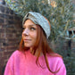 A woman with red hair wearing a textured gray and white patterned headwrap turban hat and a pink knit sweater, standing outdoors in front of a brick wall and greenery.
