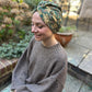 A woman wearing a green and gold floral patterned headwrap turban sits outdoors on a patio, wearing a brown knit sweater and camouflage pants.