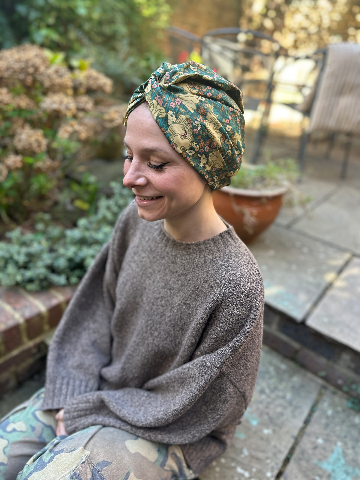 A woman wearing a green and gold floral patterned headwrap turban sits outdoors on a patio, wearing a brown knit sweater and camouflage pants.